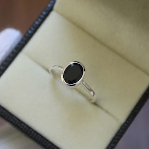 Puede incluir: Un anillo de plata con una gran piedra negra de forma ovalada, engastada en bisel. El anillo se muestra dentro de un joyero con interior beige claro.