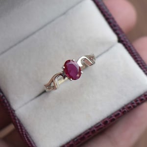 May include: A silver ring featuring an oval-cut ruby gemstone. The ruby is a deep red color and is the focal point of the ring. The band has a decorative, flowing design. The ring is presented in a maroon jewelry box.