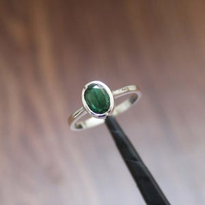 Könnte beinhalten: Ein Silberring mit einem ovalen, tiefgrünen Edelstein. Der Stein ist in einer Lünettenfassung gefasst, und das Band ist aus poliertem Silber. Der Ring wird vor einem verschwommenen Hintergrund gezeigt. Das Design des Rings ist schlicht und elegant.