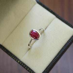 Peut inclure: Une bague en argent ornée d'un grand rubis de forme ovale. Le rubis est d'un rouge profond et est serti dans une monture à lunette. La bague est présentée dans un écrin noir avec un intérieur crème.