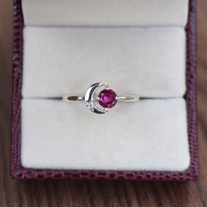 May include: A silver ring featuring a crescent moon design with a small diamond accent and a round, deep pink gemstone. The ring is presented in a burgundy jewelry box with a light interior, set against a wooden surface.