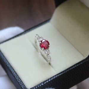 May include: A silver ring with a large, round, red gemstone as the focal point, flanked by smaller, clear gemstones. The ring is presented in a black jewelry box with a white interior, creating a striking contrast.