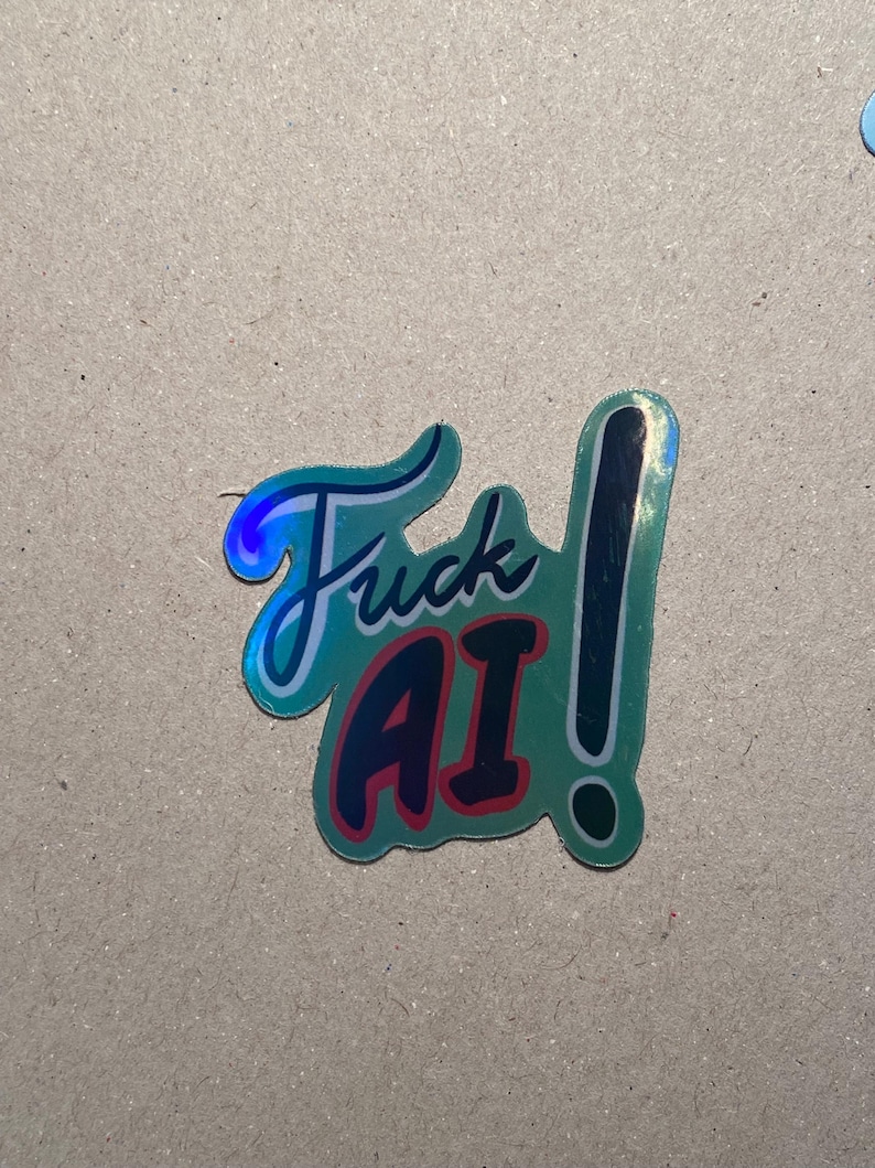May include: A teal sticker with the words "Fuck AI" in a stylized font. The word "Fuck" is in teal with a white outline, and "AI" is in red with a white outline. A black exclamation point is next to the words.