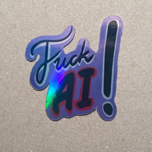 May include: A purple sticker with the words "Fuck AI" in a stylized font. The sticker has a holographic effect and a black exclamation point. The letters "AI" are in red.