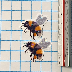 Puede incluir: Dos pegatinas de abeja con detalles en negro, naranja y blanco. Las pegatinas tienen alas iridiscentes y detalles de estrellas. Una regla con marcas de centímetros está al lado de las pegatinas, y el fondo es una cuadrícula azul.