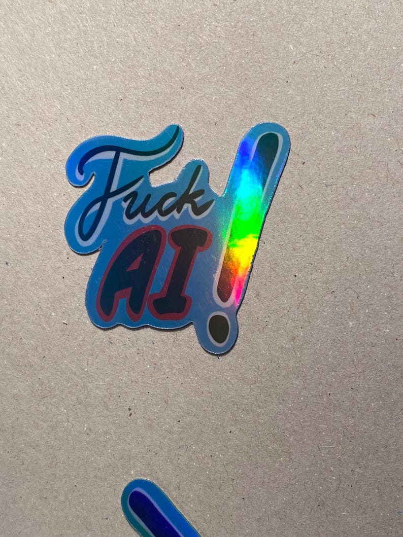 May include: A holographic sticker with the words "Fuck AI!" in a stylized font. The sticker has a blue border and a rainbow-colored exclamation point. The text "AI" is in red.