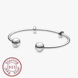 Può includere: Un braccialetto aperto in argento con tre perline decorative. Il braccialetto ha due perline sferiche alle estremità e una perlina cilindrica più piccola al centro. Il braccialetto è realizzato in argento sterling, come indicato dal timbro "Sterling 925 Silver".