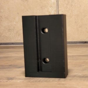 May include: A rectangular black mounting bracket featuring a recessed central channel and two circular screw holes aligned vertically. The object has a smooth, matte finish and a solid, block-like structure with clean, straight edges and corners.