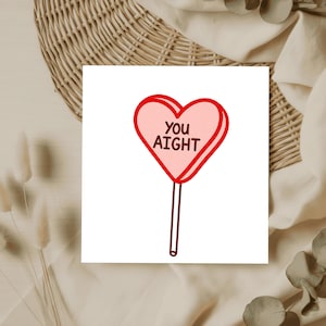 May include: A pink heart-shaped lollipop with the words "YOU AIGHT" in black text, outlined in red. The lollipop has a brown stick and is set against a white background.