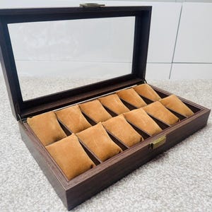 May include: A dark brown wooden watch display case with a clear glass top. The interior features twelve tan velvet cushions to hold watches. The case has a brass-colored clasp and hinges. The open lid reveals the watch storage.