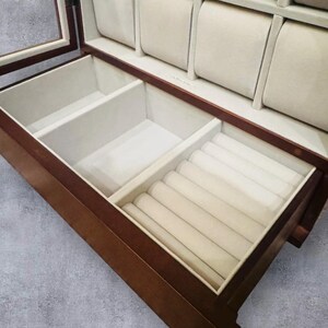 K&ouml;nnte beinhalten: Eine Holzschmuckschatulle mit Glasdeckel und mehreren F&auml;chern. Der Innenraum ist mit beigem Samt ausgekleidet und verf&uuml;gt &uuml;ber gepolsterte Uhrenhalter. Die untere Schublade hat Ringschlitze. Die Box ist aus dunkelbraunem Holz gefertigt.