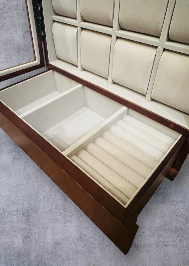 K&ouml;nnte beinhalten: Eine Holzschmuckschatulle mit Glasdeckel und mehreren F&auml;chern. Der Innenraum ist mit beigem Samt ausgekleidet und verf&uuml;gt &uuml;ber gepolsterte Uhrenhalter. Die untere Schublade hat Ringschlitze. Die Box ist aus dunkelbraunem Holz gefertigt.