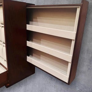 K&ouml;nnte beinhalten: Eine dunkelbraune Holzschmuckschatulle mit einer teilweise ge&ouml;ffneten Schublade, die drei cremefarbene Regale freilegt. Die Schatulle hat eine rechteckige Form und eine glatte Oberfl&auml;che mit hellem Innenraum.