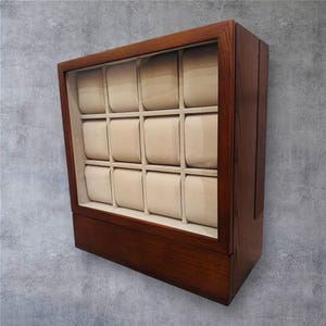 K&ouml;nnte beinhalten: Dunkelbraune Holz-Uhrenvitrine mit Glasfront. Das Geh&auml;use hat 12 beige gepolsterte F&auml;cher zur Aufbewahrung von Uhren. Der Innenraum ist mit einem hellbeigen Stoff ausgekleidet. Das Geh&auml;use ist f&uuml;r mehrere Zeitmesser konzipiert.