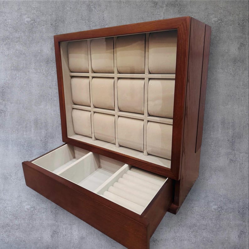 K&ouml;nnte beinhalten: Eine dunkelbraune Holz-Uhrenvitrine. Das Geh&auml;use hat einen rechteckigen Rahmen mit einem gl&auml;sernen oberen Bereich, der 12 gepolsterte, beige Uhrenf&auml;cher enth&auml;lt. Eine Schublade bietet zus&auml;tzlichen Stauraum mit F&auml;chern f&uuml;r Ringe und Accessoires.