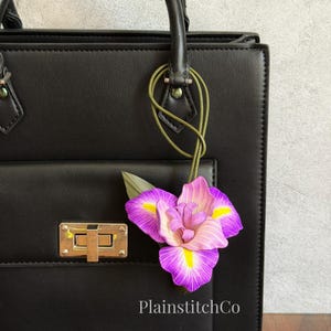 May include: A black structured handbag featuring a gold-tone rectangular turn-lock closure and a decorative purple and yellow iris flower charm attached by a thin olive green cord. The text PlainstitchCo is printed in white at the bottom.