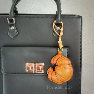 Puede incluir: Un bolso de mano de cuero negro con cierre y asa dorados. Un llavero de cuero marrón en forma de croissant cuelga del asa. El llavero está sujeto con un cordón de cuero marrón. El texto "PlainstitchCo" es visible en el bolso.