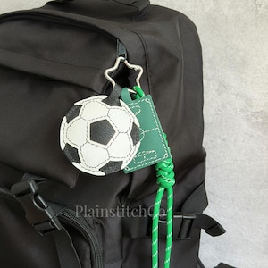Puede incluir: Una mochila negra con un llavero colgante. El accesorio incluye un dije redondo de balón de fútbol en blanco y negro, una etiqueta rectangular verde con costuras blancas y un cordón trenzado verde brillante unido a un clip en forma de estrella.
