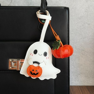 May include: A white leather ghost bag charm featuring black eyes, a small smile, and pink cheeks, holding a small orange jack-o'-lantern bucket. A separate, textured orange pumpkin charm with a green leaf is attached by a thin leather cord.