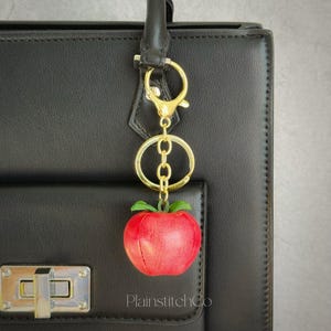 Puede incluir: Un llavero de manzana roja con hojas verdes, colgado de un cierre y una cadena dorados. El llavero está sujeto a un bolso negro. La marca "PlainstitchCo" es visible en el bolso.