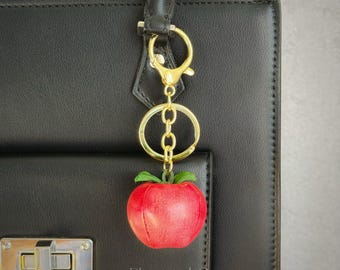 May include: A red apple keyring with green leaves, suspended from a gold-coloured clasp and chain. The keyring is attached to a black handbag. The brand name "PlainstitchCo" is visible on the bag.