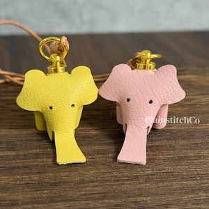 Puede incluir: Dos llaveros de elefante de cuero, uno amarillo y otro rosa. Cada uno tiene un cierre de metal dorado y un cordón de cuero. Los elefantes están sobre una superficie de madera oscura.