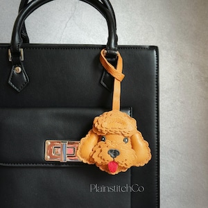 Könnte beinhalten: Ein hellbrauner Leder-Pudel-Schlüsselanhänger mit roter Zunge, der an einer schwarzen Lederhandtasche hängt. Der Schlüsselanhänger hat schwarze Augen und eine schwarze Nase. Die Handtasche hat einen silbernen Verschluss und schwarze Griffe.
