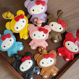 May include: A collection of colorful leather Hello Kitty-inspired keychains. Each keychain features a bow and is in various colors, including purple, yellow, blue, pink, brown, black, and red. The keychains are arranged on a wooden tray.