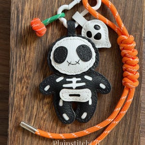 May include: A black and white faux leather skeleton character keychain featuring a white ghost charm and an orange paracord lanyard with silver metal end caps. The skeleton has stitched details and is attached to a white plastic swivel clasp.