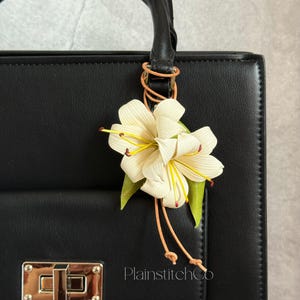 Peut inclure: Un sac à main noir avec une fermeture dorée et une poignée, orné d'un charm en forme de fleur de lys crème. La fleur a des étamines jaunes et des feuilles vertes. Le charm est attaché à la poignée avec une boucle en cuir. Le texte "PlainstitchCo" est visible.