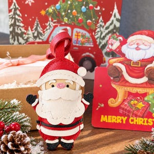 Könnte beinhalten: Ein Weihnachtsmann-Anhänger aus rotem, weißem und schwarzem Leder mit einer Schlaufe zum Aufhängen. Das Design besteht aus einem weißen Bart, einer roten Mütze mit weißem Bommel, schwarzen Handschuhen und schwarzen Stiefeln aus Leder.