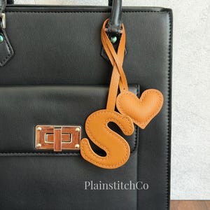 Puede incluir: Un bolso negro estructurado con un cierre giratorio dorado y un conjunto de dijes de cuero color canela que consiste en la letra S y una forma de corazón unidos al asa. El texto PlainstitchCo está impreso en blanco en la parte inferior central.