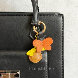 May include: A black handbag with a gold-colored ring and a charm. The charm is a colorful leather design featuring orange butterfly wings, a yellow bow, and a brown honey pot. The text "PlainstitchCo" is visible.