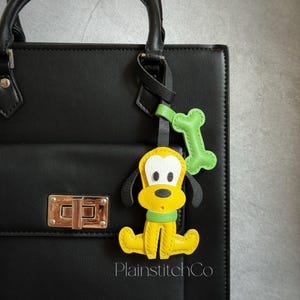 Könnte beinhalten: Ein gelber, schwarzer und weißer Hunde-Taschenanhänger mit grünem Knochenakzent. Der Anhänger ist an einer schwarzen Handtasche befestigt. Der Text "PlainstitchCo" ist auf dem Taschenanhänger zu sehen.