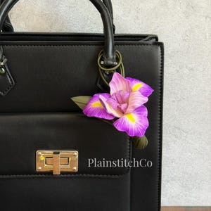 Puede incluir: Un bolso de mano negro estructurado con un cierre de giro rectangular en tono dorado y un accesorio decorativo de flor de iris morada y amarilla sujeto al asa. El texto PlainstitchCo está impreso en blanco en el bolsillo frontal.