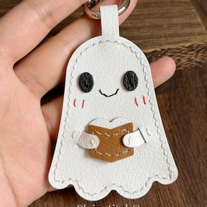 May include: A white ghost-shaped keychain featuring a textured surface, black embroidered eyes, a small black mouth, and red stitched cheeks. The ghost holds a brown book with a heart-shaped top edge. A silver metal ring is attached to the top.