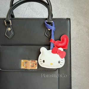 Puede incluir: Un bolso negro con cierre y asas doradas. Un charm de Hello Kitty blanco con lazo y corazón rojos está sujeto al asa con una cinta azul y roja. El texto "PlainstitchCo" es visible en el bolso.