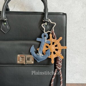 Puede incluir: Un bolso negro estructurado con un llavero de temática náutica que incluye un ancla de cuero azul, un timón de barco de cuero color canela y un cordón trenzado marrón. El llavero está unido a una anilla plateada en forma de estrella. Texto PlainstitchCo.