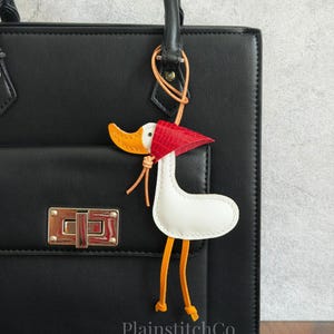 May include: A white leather goose charm with an orange beak and legs, and a red hat, hangs from a black handbag. The charm is attached to the bag with a leather strap. The handbag has a silver clasp.