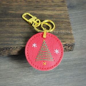 Puede incluir: Llavero redondo rojo con mosquetón y anilla dorados. El llavero presenta un diseño de árbol de Navidad bordado con copos de nieve blancos y una estrella dorada en la parte inferior. El texto "PlainstitchCo" es visible.