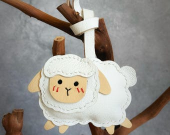 May include: A white leather sheep-shaped ornament with a cream-colored face, ears, and legs. The sheep has stitched details, black eyes, and red blush marks. The ornament is suspended from a white leather strap tied to a wooden branch. The text "PlainstitchCo" is visible.