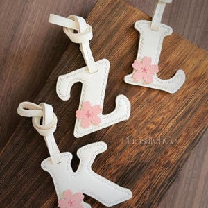 Puede incluir: Tres etiquetas de equipaje de cuero blanco con forma de letras "Z", "L" y "K", cada una con un acento de flor rosa. Las etiquetas tienen una correa anudada y están sobre una superficie de madera. La marca "PlainstitchCo" es visible.