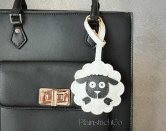 May include: A black leather handbag with a gold clasp and handles. A white and black sheep-shaped keychain is attached to the handle. The keychain has a fluffy white body and a black face. The text "PlainstitchCo" is visible.