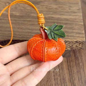 Könnte beinhalten: Oranger Leder-Kürbis-Anhänger mit grünem Blatt und Lederband. Der Kürbis ist mit orangefarbenem Faden genäht. Ein Accessoire zum Thema Herbst.