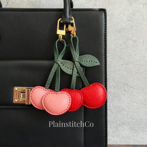 Puede incluir: Un colgante de cuero para bolso con forma de cerezas que presenta dos pares de cerezas en tonos rosa claro y rojo brillante, unidas a tallos y hojas verdes. El colgante está sujeto con un cierre de metal dorado en un bolso negro con el texto PlainstitchCo.
