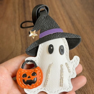 May include: A white ghost-shaped leather keychain featuring a black witch hat with a purple band and a small star charm. The ghost holds an orange pumpkin bucket with black facial features and has silver beaded accents on its body.