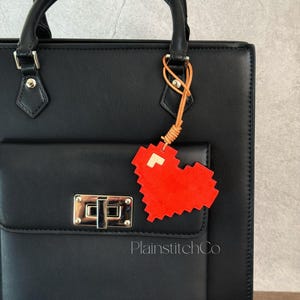 Puede incluir: Bolso tote de cuero negro con solapa delantera y cierre plateado. Un charm de corazón pixelado rojo cuelga del asa. El charm está sujeto con un cordón de cuero marrón claro. El texto "PlainstitchCo" es visible en el bolso.