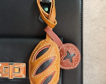 Puede incluir: Un colgante de cuero para bolso con forma de pan ovalado de color tostado, con detalles en marrón oscuro y costuras blancas, sujeto a un bolso negro. Una etiqueta de cuero circular más pequeña con una estrella recortada y costuras cuelga al lado.