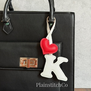 May include: A black handbag featuring a gold-tone metal clasp and a hanging charm. The charm consists of a red heart and a white letter K, both crafted from a textured material with visible stitching, suspended by a white strap from the handle.