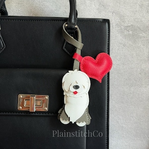 Puede incluir: Un bolso de cuero negro con un cierre plateado, con un charm de perro de cuero hecho a mano y un charm de corazón rojo. El charm de perro es blanco y gris, con una lengua roja. El texto "PlainstitchCo" es visible.
